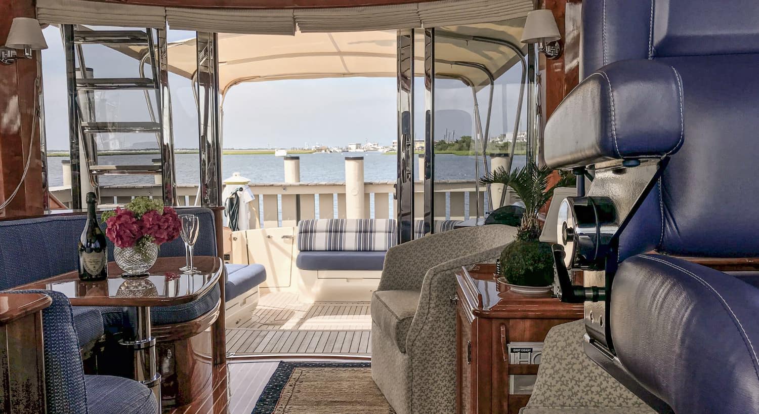 Inside of motor sailing yacht with blue cushioned sitting area, wooden table, upholstered armchair, and view of the back covered sitting area