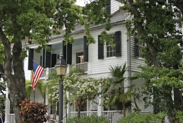 Southern style home painted white with dark green shutters, wrap around porch, and surrounded by green trees and bushes